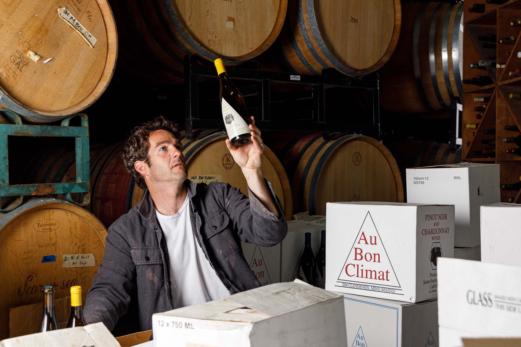 A man examining a bottle that he's holding up above an Au Bon Climat branded case box with wine barrels behind.