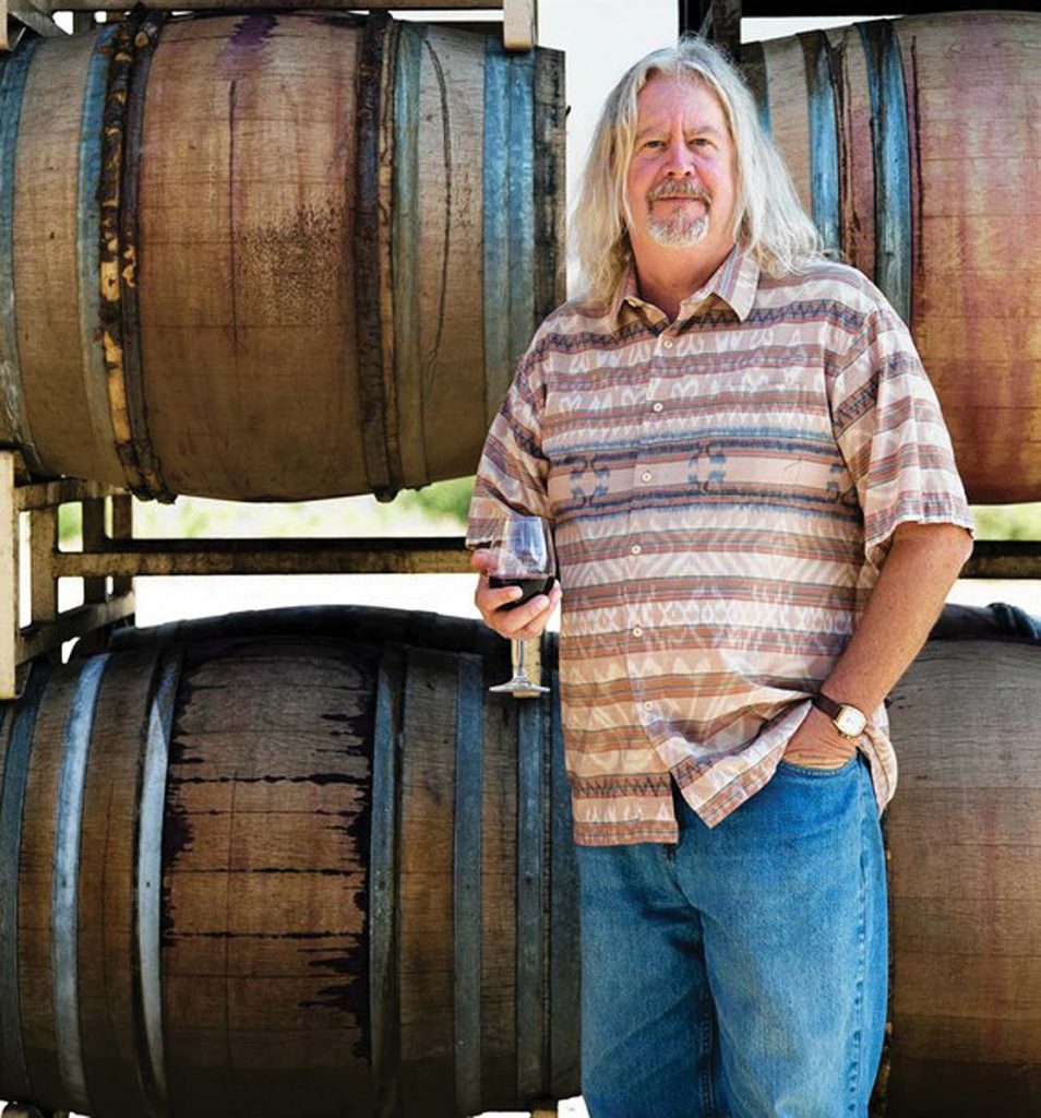 Jim Clendenen standing in front of wine barrels holding a glass of wine.